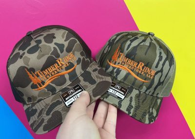 Two camo trucker hats embroidered with an orange Timber Ridge logo by 400 Ink